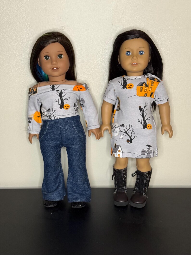 May include: Two dolls wearing Halloween-themed outfits. One doll is wearing a gray off-the-shoulder top with a pumpkin and tree print, paired with blue denim bell-bottom pants. The other doll is wearing a gray dress with a similar print and black boots.