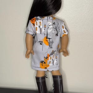 May include: A doll wearing a gray dress with a Halloween-themed print featuring orange pumpkins, houses, and a full moon. The doll has long black hair and is wearing brown boots.