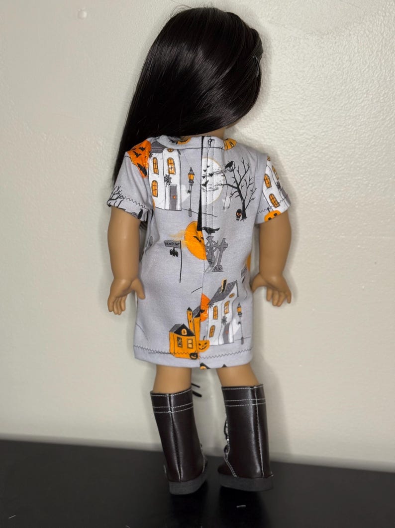 May include: A doll wearing a gray dress with a Halloween-themed print featuring orange pumpkins, houses, and a full moon. The doll has long black hair and is wearing brown boots.