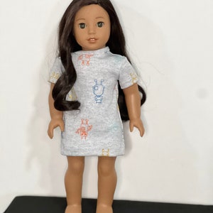 May include: A doll with long dark hair wearing a light gray dress. The dress features a pattern of colorful robot illustrations. The doll is standing on a black surface against a white background.