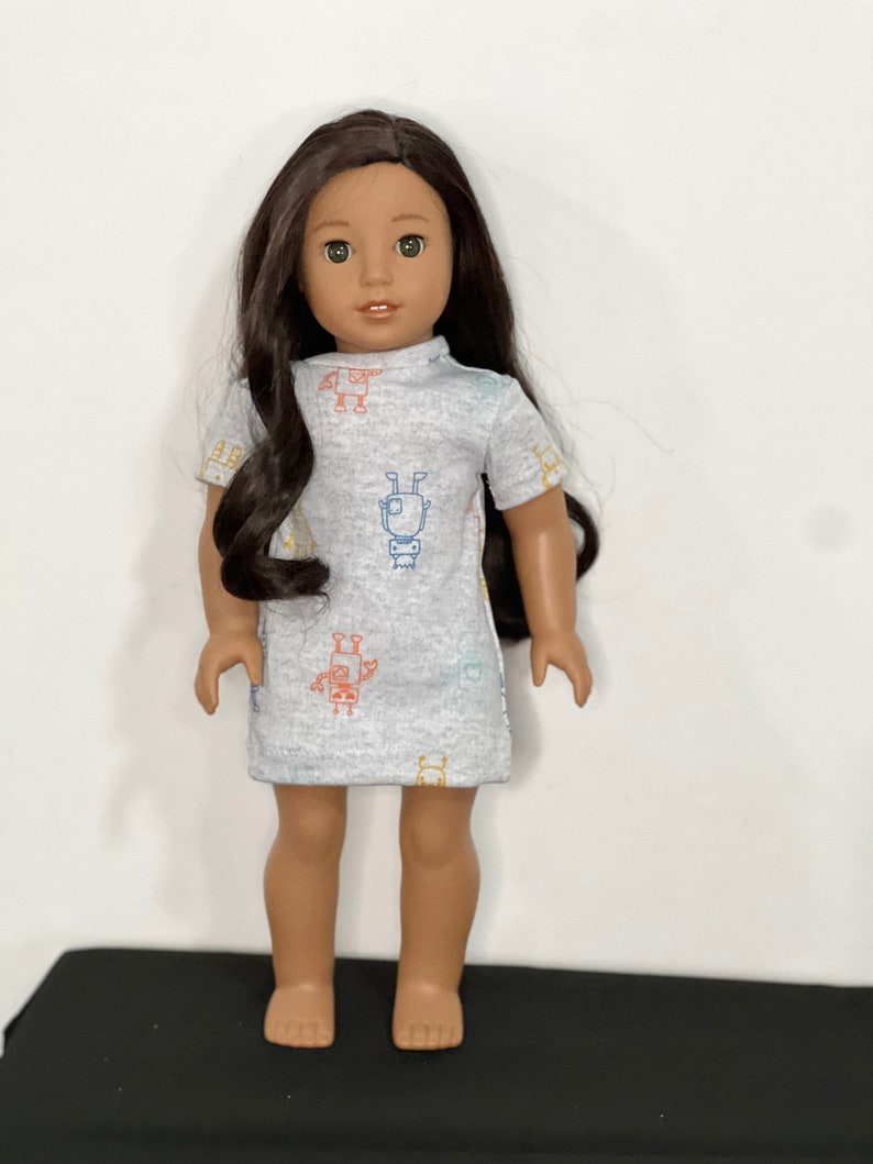 May include: A doll with long dark hair wearing a light gray dress. The dress features a pattern of colorful robot illustrations. The doll is standing on a black surface against a white background.