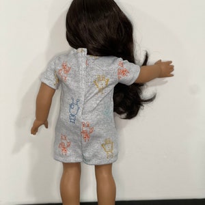 May include: A doll wearing a gray romper with a colorful robot pattern. The romper has short sleeves and a button closure on the back. The doll has long, dark brown hair.