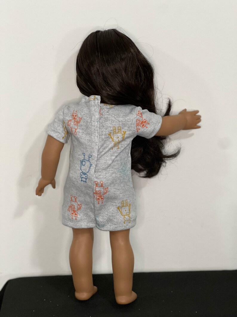 May include: A doll wearing a gray romper with a colorful robot pattern. The romper has short sleeves and a button closure on the back. The doll has long, dark brown hair.