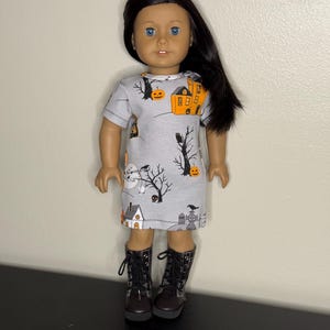 May include: A doll wearing a gray Halloween-themed dress with orange houses, pumpkins, and black trees. The doll has black boots and long black hair. The dress is short-sleeved.