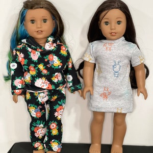 May include: Two dolls are shown. One doll wears a black floral print hoodie and matching pants. The other doll wears a gray dress with a robot pattern. Both dolls have long brown hair and are standing.