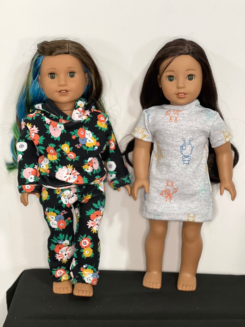 May include: Two dolls are shown. One doll wears a black floral print hoodie and matching pants. The other doll wears a gray dress with a robot pattern. Both dolls have long brown hair and are standing.