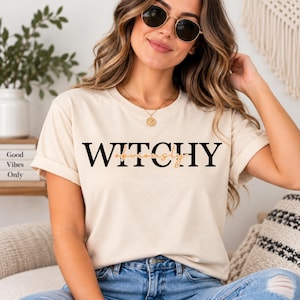 May include: A cream-colored short-sleeve t-shirt featuring the word WITCHY in bold black capital letters with the word obviously written in a smaller, cursive orange font layered across the center of the text on the front of the garment.