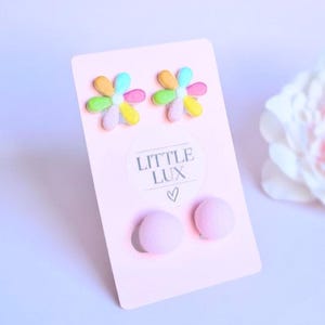 May include: A pair of flower-shaped earrings with pastel petals and a pair of pink ball earrings are displayed on a pink card. The card has the text "LITTLE LUX" and a heart symbol.