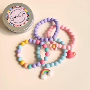 May include: A creative bracelet kit in a round metal tin labeled Bead and Create, accompanied by three elastic bracelets made of matte pastel beads. The bracelets feature charms including a pink flower, a red heart, and a colorful rainbow.
