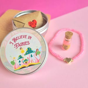 May include: A pink beaded bracelet with a gold heart charm and a small glass bottle filled with pink glitter. A silver tin with the words "I Believe in Fairies" and a fairy illustration is open, revealing a small envelope with a red heart.