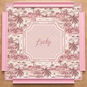 May include: A pink board game with a tropical landscape design featuring palm trees, boats, and buildings. The center has a geometric pattern with the word "Lucky". The board is framed by pink edges and includes game instructions.