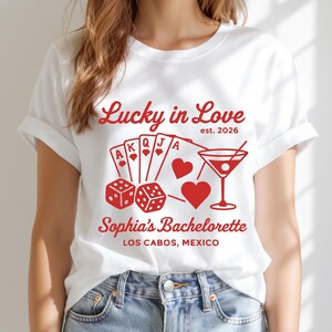 May include: A white short-sleeve t-shirt featuring red graphic text and illustrations. The design includes the text Lucky in Love est. 2026, playing cards, two dice, two heart shapes, a martini glass, and the text Sophia's Bachelorette LOS CABOS, MEXICO.