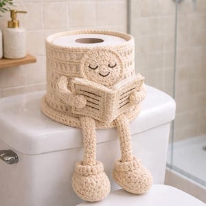 May include: A beige crochet toilet paper cover shaped like a cylinder, with a smiling face and legs. The cover is reading a book and sitting on a white toilet. The bathroom has a neutral color scheme.