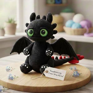 Könnte beinhalten: Ein handgefertigter, schwarzer Häkeldrachen mit großen grünen Augen, Flügeln und einer roten Schwanzspitze. Der Drache sitzt auf einer Holzoberfläche, mit einem Anhänger mit der Aufschrift "Handmade by [Your Shop Name] ". Ein niedliches, skurriles Spielzeug.
