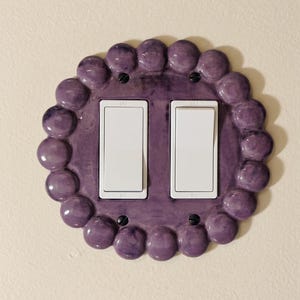 May include: A double light switch wall plate featuring a glossy purple ceramic border with a raised, scalloped, bead-like edge. The center holds two rectangular white rocker switches, each labeled with ON and OFF text, secured by four black screws.