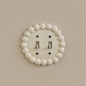 May include: A circular, off-white light switch plate featuring a decorative scalloped edge made of rounded, bead-like protrusions. The plate accommodates two toggle switches and is secured by four small, dark-colored screws on the face.