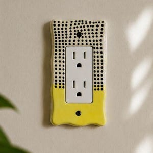 Peut inclure: Une plaque murale d'interrupteur en céramique rectangulaire avec un bord ondulé. La partie supérieure est de couleur crème avec de petits points noirs, tandis que la partie inférieure est d'un jaune vif uni. Une prise électrique blanche est centrée.