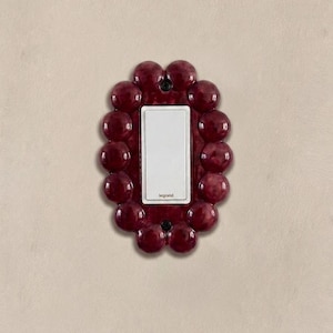 May include: A rectangular light switch with a white center plate featuring the brand name legrand in small black text. The switch is surrounded by a decorative frame made of glossy, dark red, spherical ceramic-like orbs arranged in a cluster.