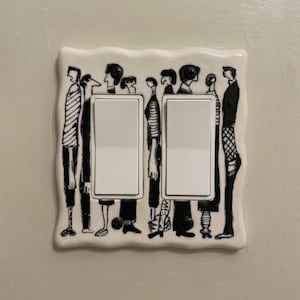 May include: A white double light switch wall plate with a wavy edge, featuring a black and white line art illustration of a crowd of people standing side by side. The design surrounds two rectangular white rocker switches.