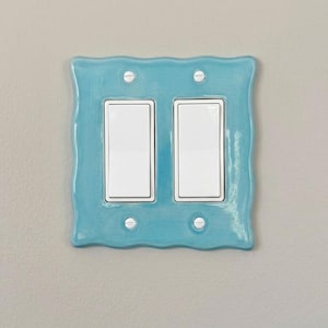 Peut inclure: Une plaque murale pour double interrupteur avec un cadre en céramique turquoise brillant aux bords ondulés. La plaque comporte deux interrupteurs à bascule blancs rectangulaires centrés, fixés par quatre petites vis métalliques argentées.