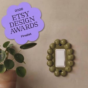 May include: A light switch cover plate featuring a glossy olive green ceramic frame with a series of rounded, bubble-like protrusions surrounding a central white rectangular switch. A purple cloud-shaped badge displays 2026 ETSY DESIGN AWARDS Finalist.
