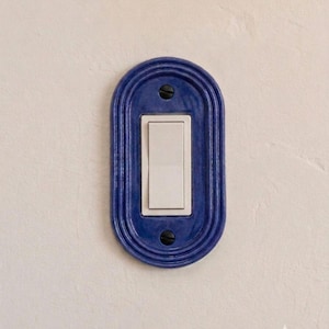 May include: An oval-shaped, deep blue ceramic light switch plate featuring a glossy finish and concentric ridged detailing. The plate surrounds a standard white rectangular rocker light switch, secured by two visible black screws at the top and bottom.