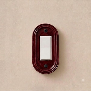 May include: An oval-shaped, deep red wall plate featuring a glossy finish and concentric ridged detailing. A rectangular white light switch is centered within the plate, secured by two small, dark-colored screws at the top and bottom positions.