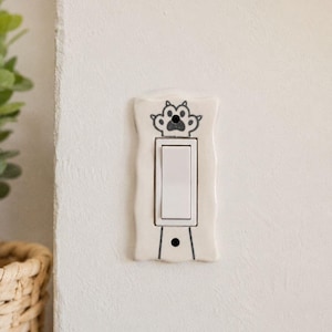 May include: A rectangular off-white ceramic light switch cover plate featuring a hand-drawn black outline of a cat paw print at the top and a single line extending downward. The plate has a wavy edge and a central rectangular cutout for a rocker switch.