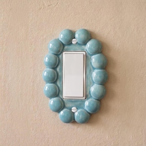 May include: A light blue ceramic light switch cover plate featuring a decorative border of rounded, bubble-like spheres. The rectangular white rocker switch is centered within the oval frame, secured by two visible silver-toned metal screws.