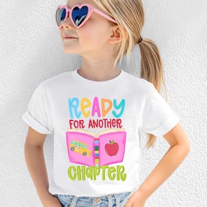 Puede incluir: Una camiseta blanca de manga corta con un gráfico colorido de un libro rosa abierto que muestra un autobús escolar amarillo y una manzana roja. El texto Ready For Another Chapter aparece en letras brillantes de color azul, naranja, rojo y verde.
