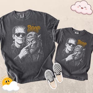 May include: Two charcoal gray t-shirts featuring a graphic of Frankenstein's monster wearing sunglasses and a werewolf wearing a baseball cap. The monster is poking the werewolf's nose, with the word Boop printed in gold above them.