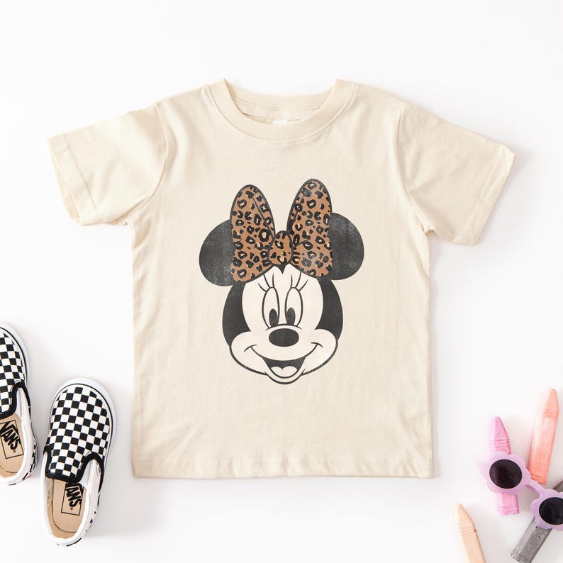 May include: Cream-colored t-shirt featuring a distressed graphic of Minnie Mouse's face. The bow has a leopard print design. The shirt is displayed with black and white checkered slip-on shoes, sunglasses, and crayons.