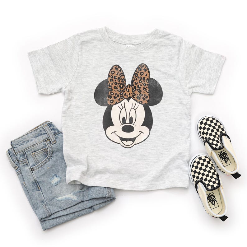 May include: A light gray t-shirt featuring a Minnie Mouse graphic with a leopard print bow. The image also includes a pair of light blue denim shorts and black and white checkered slip-on shoes.