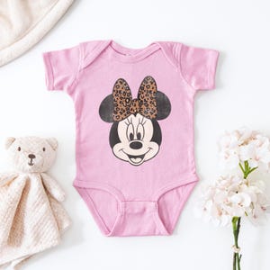 May include: A light pink baby onesie featuring a Minnie Mouse graphic. The Minnie Mouse design has a black face, ears, and a bow with a leopard print pattern. A teddy bear and a white blanket are in the background.