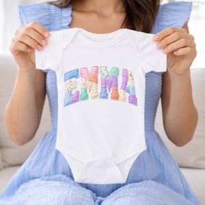 May include: White baby onesie with the name "EMMA" appliqued in colorful patchwork fabric. The letters are in shades of blue, pink, green, and purple. The onesie is short-sleeved and has a snap closure.