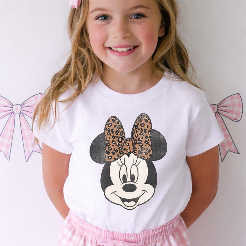 May include: White t-shirt featuring a Minnie Mouse graphic with a leopard print bow. The graphic is centered on the shirt. The girl is wearing pink gingham shorts and a pink bow in her hair.