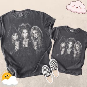 May include: Two charcoal gray t-shirts featuring a black and white graphic print of three women holding clear plastic cups with straws. The print includes the names Mary Sanderson, Winifred Sanderson, and Sarah Sanderson on the cups.