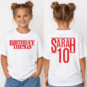 May include: White t-shirt with red text that says "BIRTHDAY THINGS" on the front and "SARAH 10" on the back. The shirt is short-sleeved.