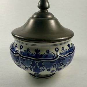 Peut inclure: Un pot en céramique avec un couvercle gris foncé et un motif floral bleu et blanc. Le pot a une forme arrondie et repose sur une petite base. Le couvercle a un petit bouton arrondi sur le dessus. Le motif floral est répété.
