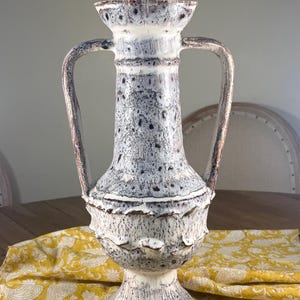 Peut inclure: Un vase en céramique tacheté et de grande taille, avec deux anses. Le vase présente un motif de couleurs blanc, gris et marron avec une surface texturée. Le vase est posé sur une table en bois avec un tissu jaune à motifs.