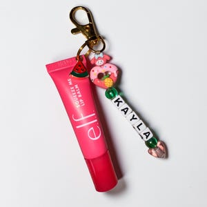 May include: A pink e.l.f. Squeeze Me lip balm tube with a gold keychain, a watermelon charm, and a charm shaped like a donut with a character. A beaded charm spells out "KAYLA".