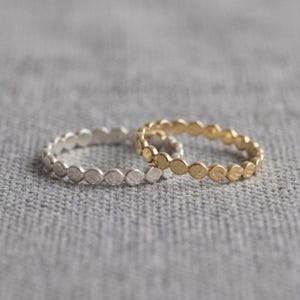 Könnte beinhalten: Zwei Ringe, einer silberfarben und einer goldfarben, jeweils mit einer Reihe kleiner, gleichmäßig verteilter Kreise verziert. Die Ringe liegen auf einer strukturierten, grauen Oberfläche und bilden eine schlichte, aber elegante Komposition. Der goldene Ring hat ein warmes, glänzendes Finish.