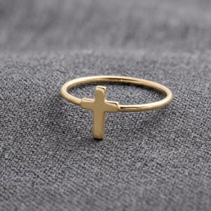 May include: A gold ring with a cross design. The ring has a slender band and a simple cross at the centre. The ring is set against a grey textured background.