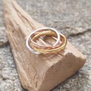 Può includere: Un anello a tre fasce intrecciate nei toni dell'argento, dell'oro e dell'oro rosa. Ogni fascia presenta una superficie martellata e testurizzata che cattura la luce, creando un aspetto stratificato con una finitura lucida.