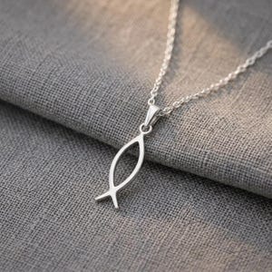 Może przedstawiać: Srebrny naszyjnik z zawieszką w kształcie chrześcijańskiego symbolu ryby. Zawieszka ma prosty, otwarty design, zawieszona na delikatnym łańcuszku. Naszyjnik spoczywa na tle szarej, teksturowanej tkaniny.