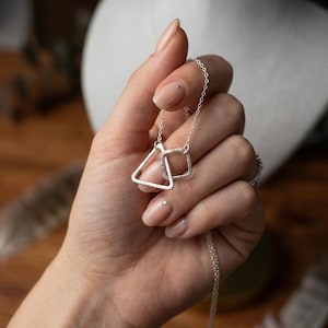 May include: A silver necklace featuring a geometric design. The pendant includes a triangle and a square, connected by a delicate chain. The jewelry is displayed against a neutral background, highlighting the minimalist style.