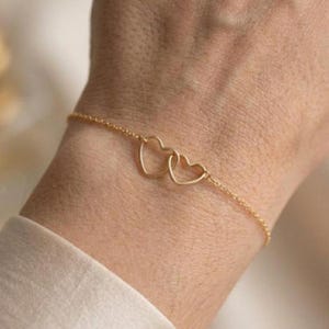 May include: A delicate gold bracelet featuring two intertwined heart-shaped charms. The bracelet is a fine chain, and the hearts are open, creating a minimalist and elegant design. The bracelet is worn on a wrist.