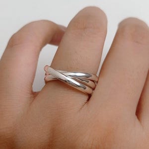 Pode incluir: Um anel prateado com um design de três bandas. As bandas lisas se cruzam, criando um padrão entrelaçado. O anel é usado em um dedo, refletindo a luz. Joalharia.