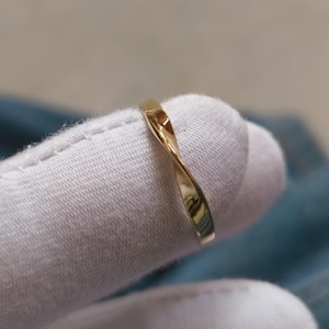 May include: A gold-colored ring with a twisted design. The ring is smooth and reflective, catching the light. The ring is shown on a white surface.