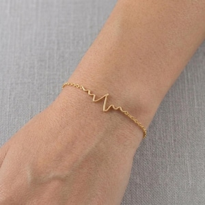 Op de afbeelding: Een delicate goudkleurige armband met een hartslagontwerp. De armband is een dunne ketting met een gestileerd elektrocardiogrampatroon. De sieraden worden om de pols gedragen, wat wijst op een thema van gezondheid of medische symboliek.